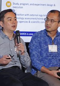 One man speaks into a microphone while another man looks on
